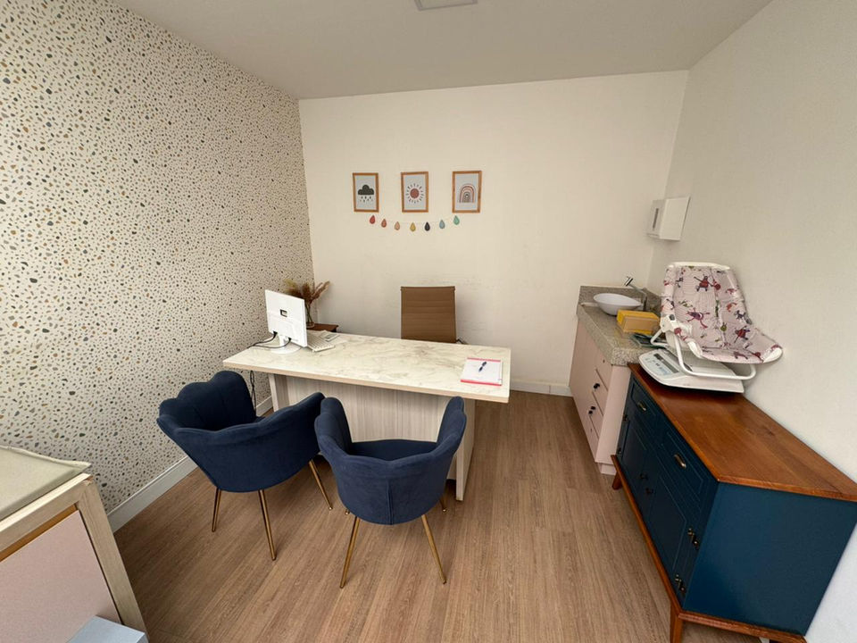 Sala de atendimento com mesa de consulta, poltronas e balança pediátrica